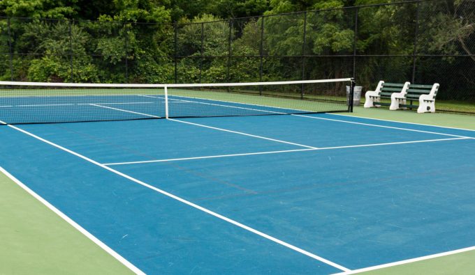 A blue tennis court inside a black fence with be3nches in the middle surrounded by tall trees in the background.
