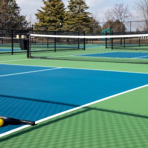 View of a pickleball complex with a paddle and yellow on blue and green courts beside a playground in a suburban park in early spring.